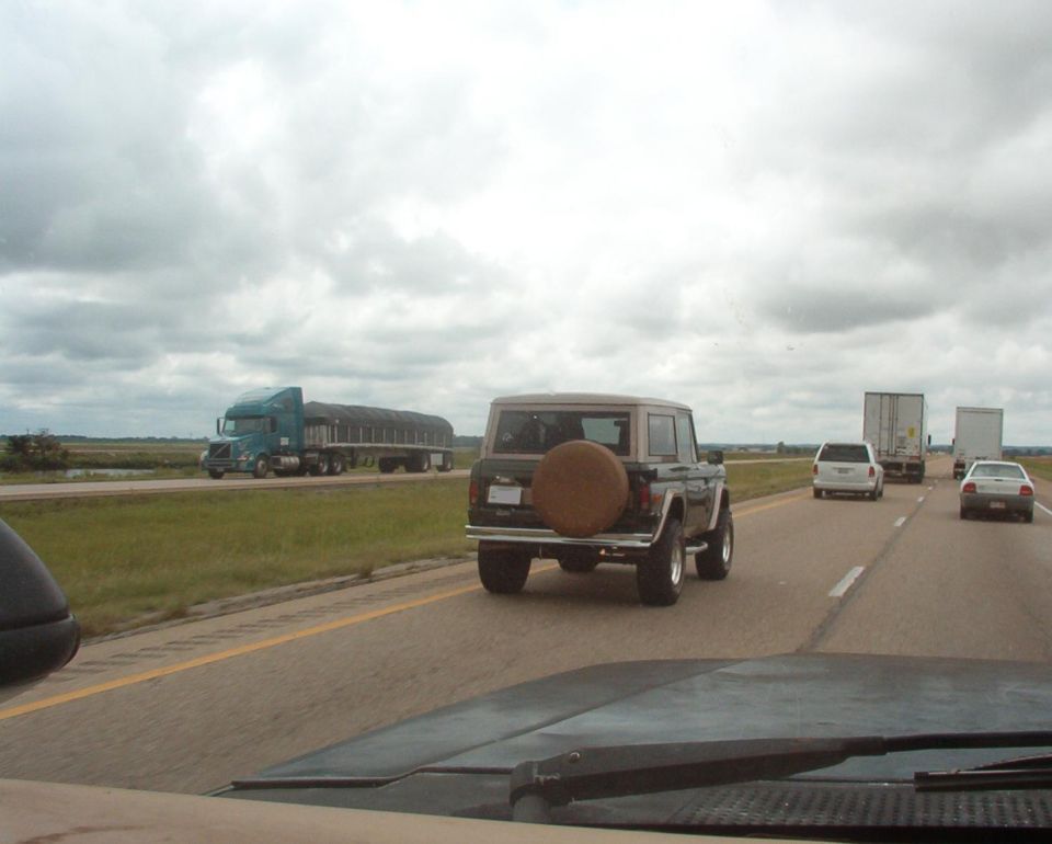 Dark green early Ford Bronco with white top and covered rear spare on a highway