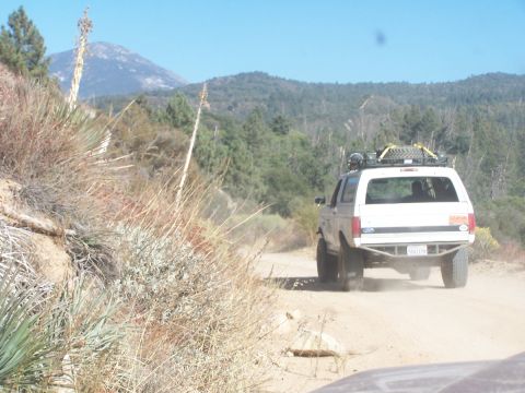 1990 Ford Bronco - A white 1990 Ford Bronco kicks up dust as it travels along a remote dirt road, with mountains and dry vegetation in the background.