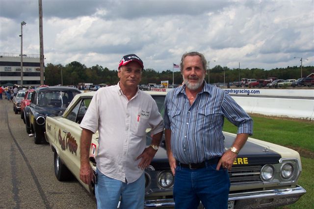 Two men stand by a 1967 Plymouth GTX at a drag strip. Other cars are lined up behind it