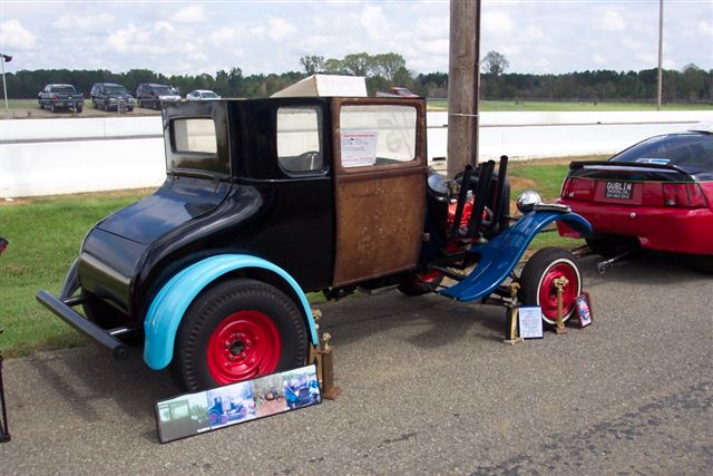 Modified hot rod with black body, blue fenders, and exposed engine. Red Mustang in background