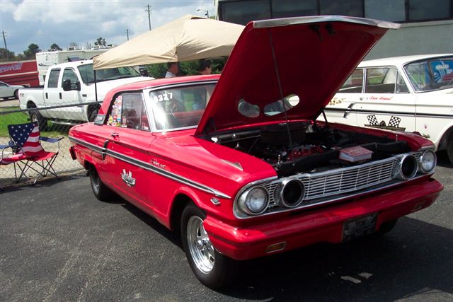 Red 1963 Ford Fairlane coupe with hood open, showing engine. White Ford truck and another car in background