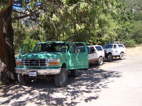 1990 Ford Bronco - Three-quarter front view of a green 1990s Ford F-Series 4x4 crew cab with both driver-side doors open, parked in tree shade on a dirt turno…
