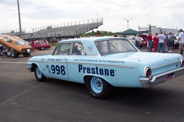 light blue race car with Prestone sponsorship and number 998, parked at a drag strip with bleachers in the background