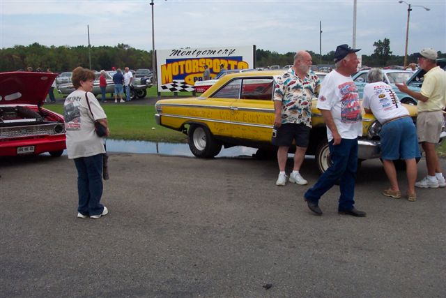 Yellow 1964 Ford sedan parked at an outdoor event with several people standing around it. A red car with its hood open is visible to the left