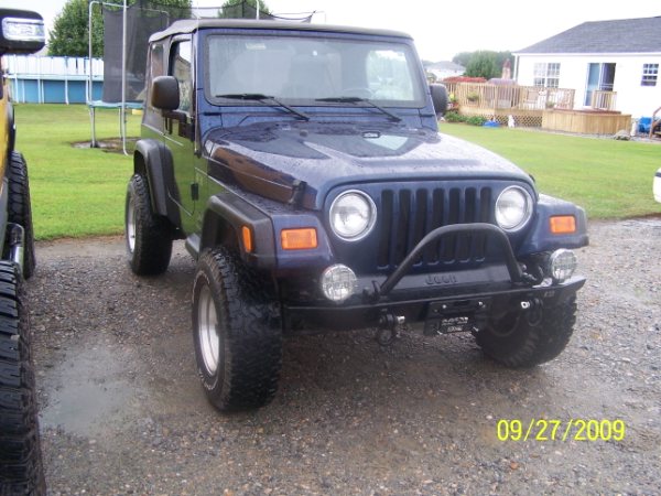 Dark blue 2003 Jeep Wrangler with a bull bar and aftermarket lights parked on gravel