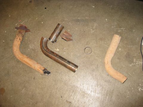 1995 Ford Bronco - Three sections of exhaust tubing laid out on a concrete shop floor: a rusty crushed-end downpipe with a flaking flange, a freshly cut and b…