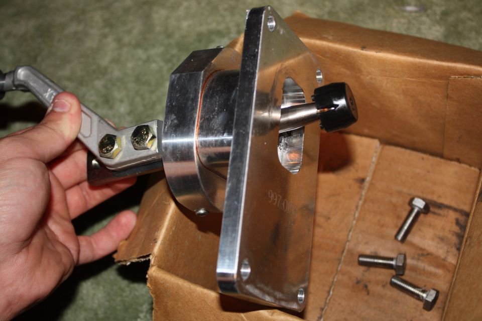 Hand holding metal shifter assembly with black knob, next to bolts in cardboard box