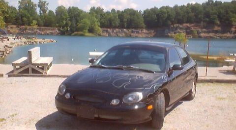 1999 Ford Taurus - A black sedan with flame decals sits parked near a body of water with trees in the background.