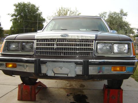 1984 Ford LTD - Front view of a brown Ford car with a chrome grille and dual headlights, raised on red jack stands.