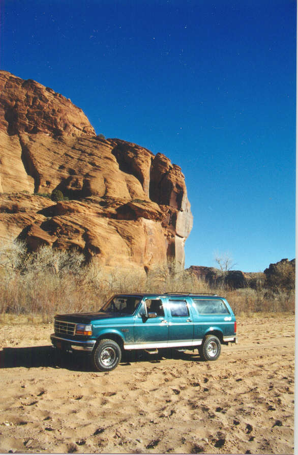 Teal four-door Ford Centurion SUV parked on sand in front of red sandstone cliffs