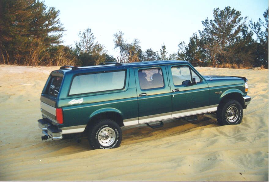 Green four-door Centurion Ford SUV conversion parked on sand dunes, rear three-quarter view, white-letter tires