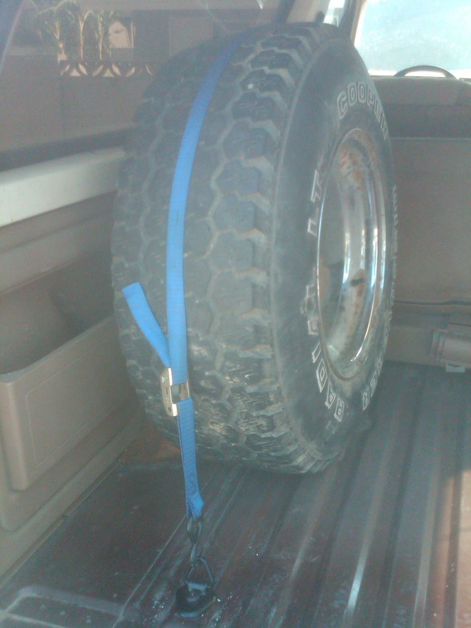 Spare tire with Goodyear Radial text, secured by a blue strap in a vehicle's cargo area