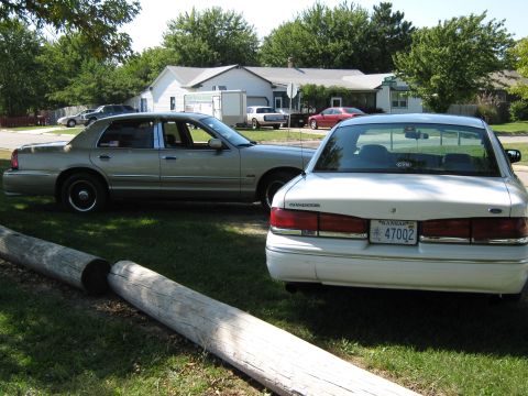2000 Mercury Grand Marquis - Two sedans are parked on a grassy area in front of houses, one is a white sedan with its rear facing the camera.
