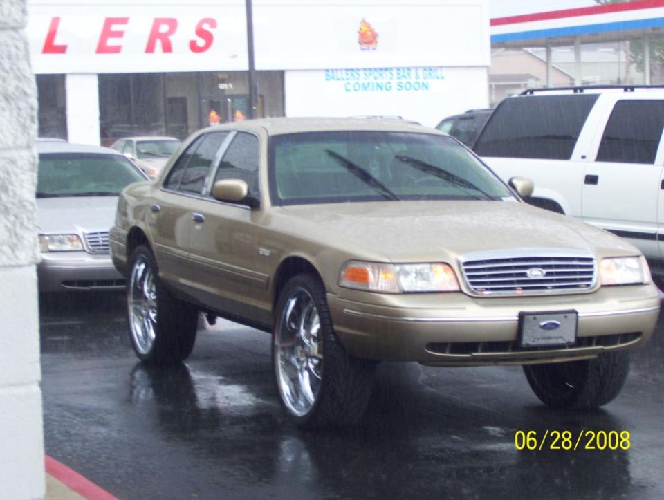 Gold Ford Crown Victoria sedan on large chrome wheels, parked on wet pavement.