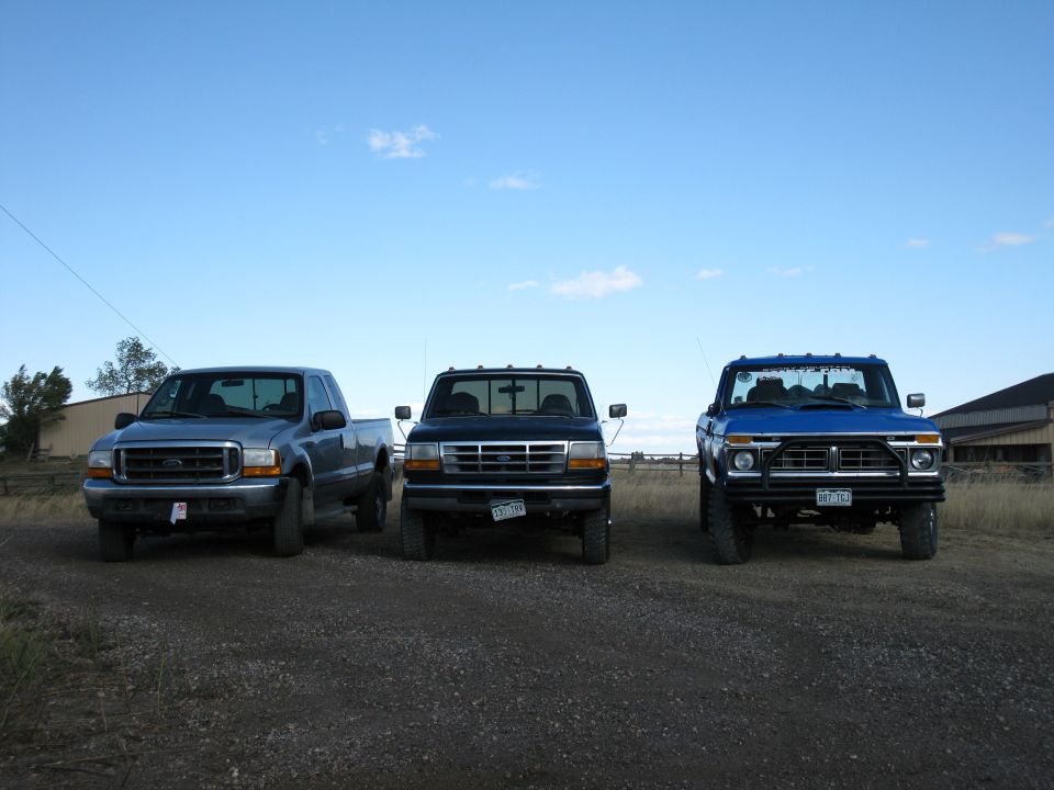 Three pickup trucks parked in a line on a dirt road. The truck on the left is blue, the middle truck is black, and the truck on the right is blue