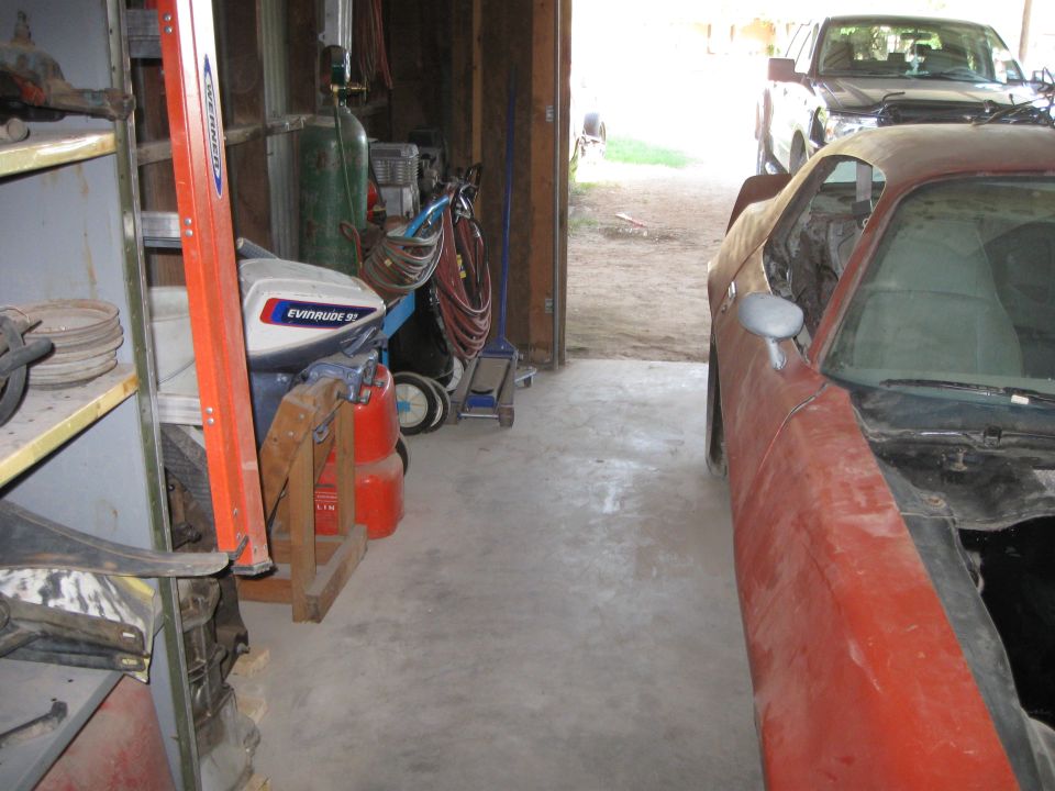 Red car body in garage next to a pickup truck. Tools and an outboard motor are also visible