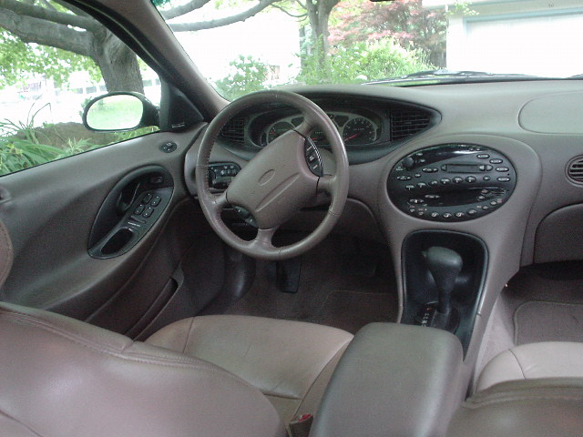 Beige sedan interior dash and steering wheel with automatic shifter and Ford oval on horn pad