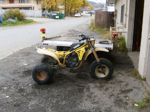 2000 Ford E350 - A yellow Yamaha three-wheeler with a flat front tire is parked on a trailer next to a building.