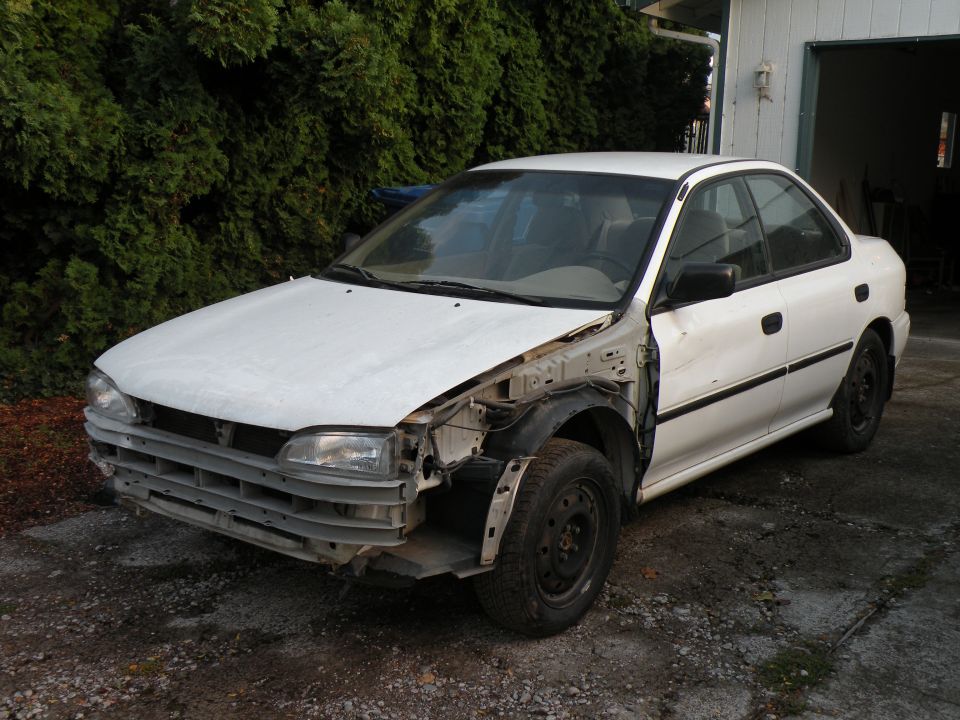 White sedan with front end damage, missing grille and bumper. Visible fender, headlight, and tire