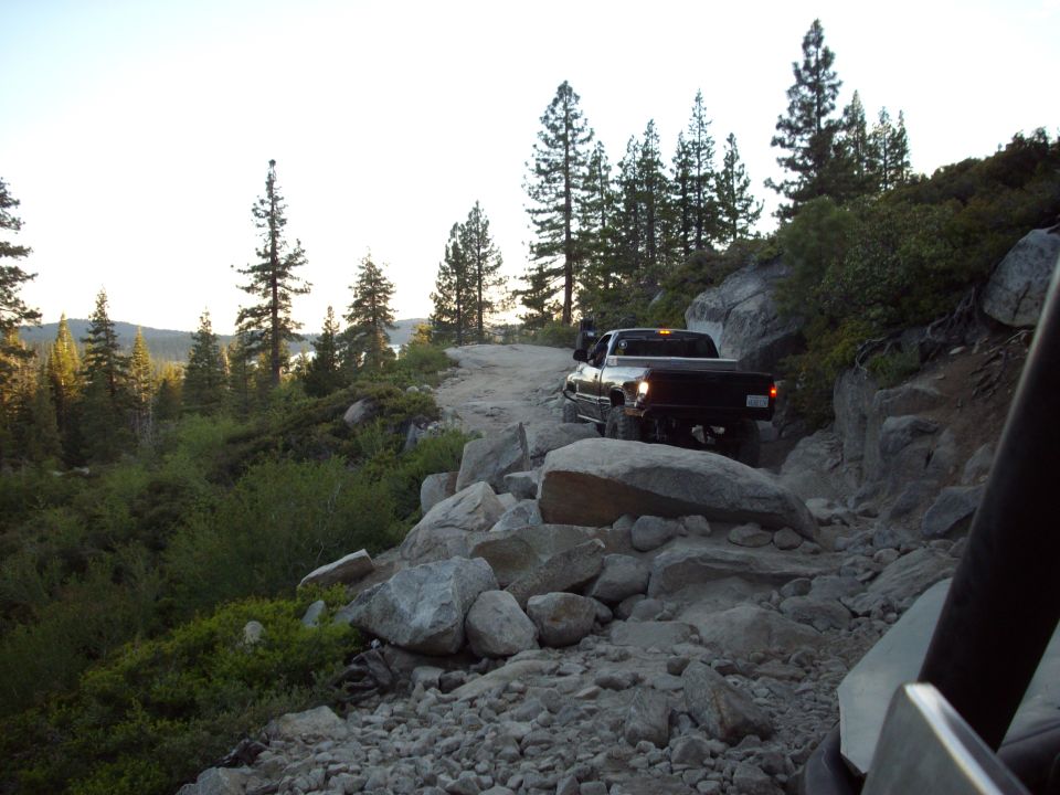 Pickup truck navigating a rough, rocky trail with trees and a lake in the background