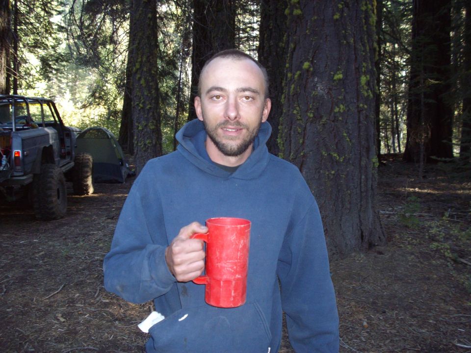 Man holding a red mug in a forest campsite with a modified off-road vehicle and tent in the background