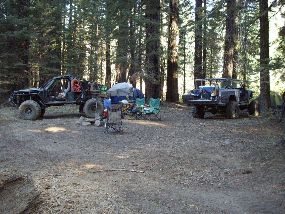 Two modified off-road vehicles parked at a campsite in a forest. One vehicle is a tube-frame buggy, the other is a pickup truck with camping gear in the bed