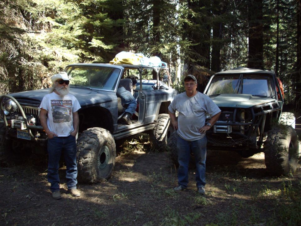 Two guys stand next to heavily modified off-road vehicles in a wooded area