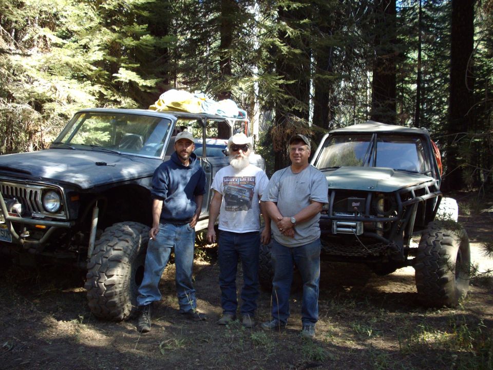 Two lifted trail rigs parked in a pine forest with three men standing between them