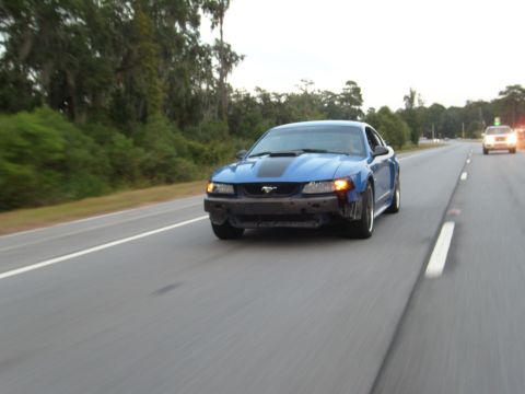 1992 Ford Bronco - Blue Ford Mustang with a missing front bumper drives down the highway, with another vehicle visible ahead.