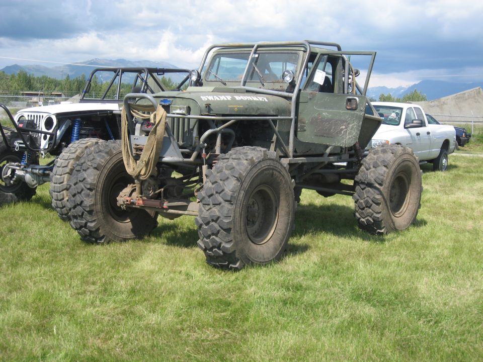 Custom off-road jeep with large tires and roll cage, labeled "SWAMP DONKEY", parked next to another modified jeep and a white pickup truck