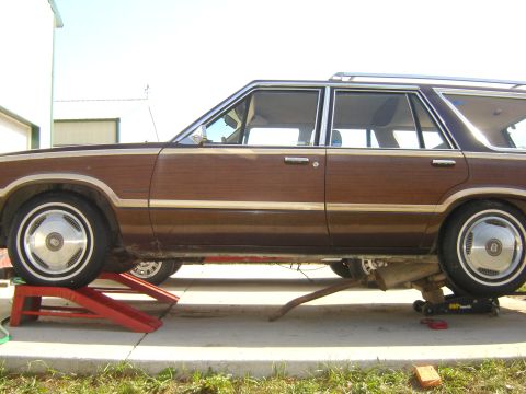 1984 Ford LTD - A brown station wagon with wood paneling is elevated on red ramps and a jack.