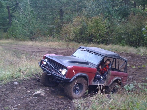 Red 1968 Ford Bronco with black hood and soft top, tilted on a muddy trail with a driver giving a thumbs up