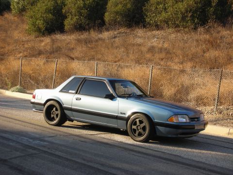 1990 Ford Mustang - A light blue 1990 Ford Mustang is parked on the side of a road with tire marks visible on the asphalt.