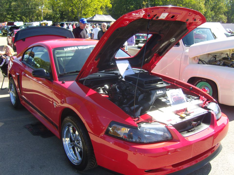 Red Mustang with hood open, showing engine. Another classic car is parked next to it