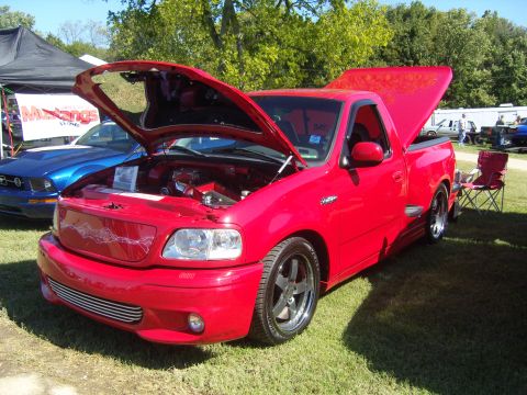 2007 Ford Mustang - A red pickup truck is displayed with its hood open, parked next to a blue Ford Mustang at an outdoor event.