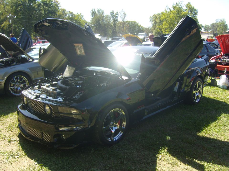Black 2007 Ford Mustang with hood and lambo doors open, parked on grass at an outdoor car show