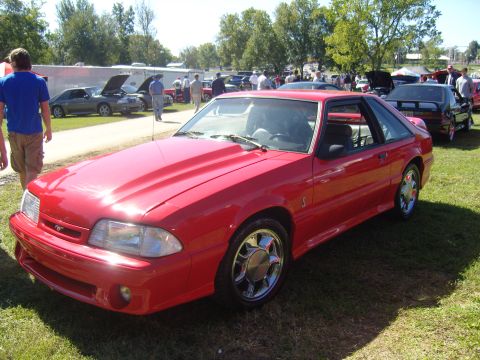 2007 Ford Mustang - A red coupe with a hood scoop is parked on the grass at an outdoor event.