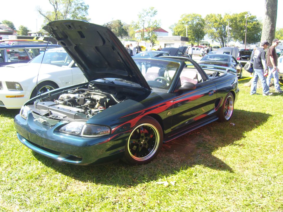 Dark green convertible Mustang with racing stripes, hood open showing engine. Another white Mustang is parked nearby