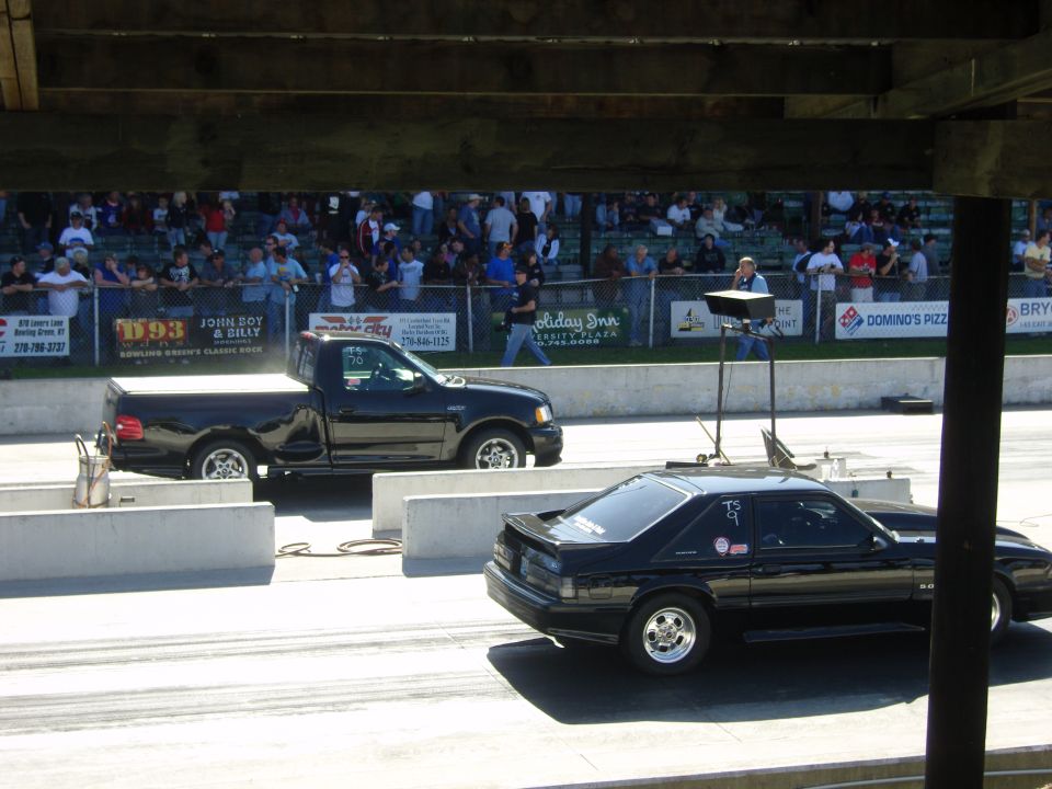 Black pickup truck and black coupe drag racing on a track with spectators in the background