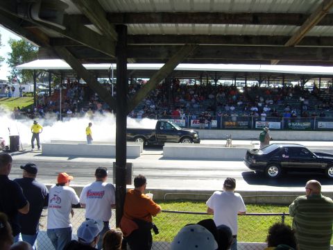 2007 Ford Mustang - Two cars perform burnouts on a drag strip under a covered spectator area.