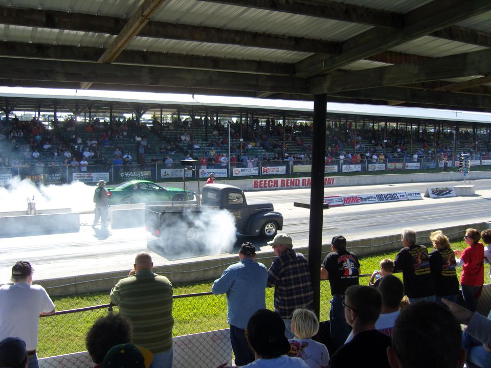 Black pickup truck doing a burnout at a drag strip, with a green car in the background