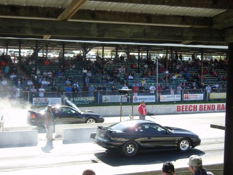 2007 Ford Mustang - Two Mustangs compete in a drag race, with spectators in the stands and smoke billowing from the tires.