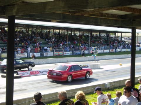 2007 Ford Mustang - A red Ford Mustang competes on a drag strip, with spectators in the stands watching the race.