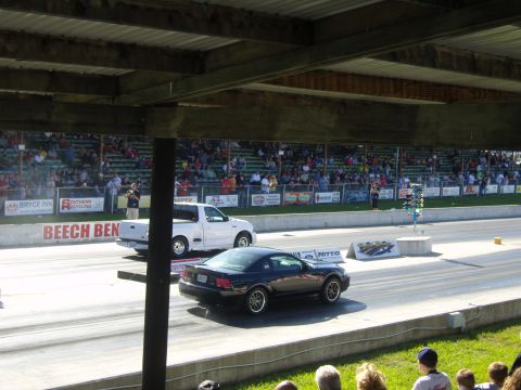2007 Ford Mustang - A black Ford Mustang and a white pickup truck compete on a drag strip, with a crowd watching from the bleachers.