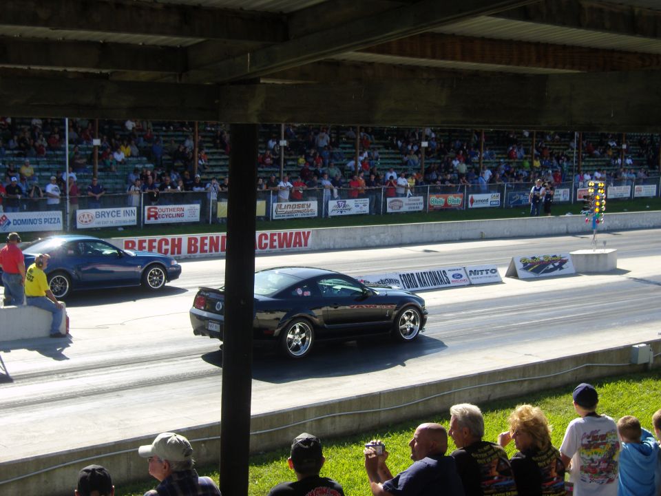 Black Ford Mustang racing on a drag strip, blue Mustang in background. Crowd watching from bleachers