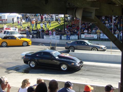 2007 Ford Mustang - Two Ford Mustangs compete on a drag strip, with spectators watching from the stands.