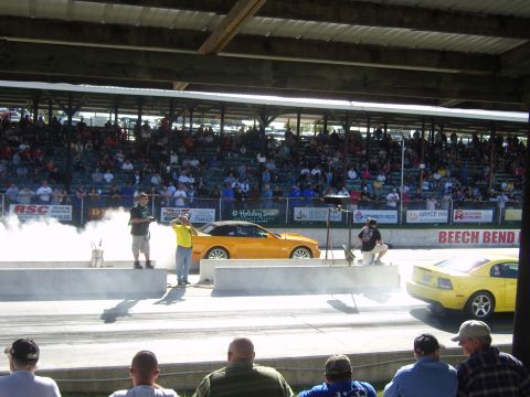 2007 Ford Mustang - Two yellow cars are on a drag strip with a crowd watching from the stands.