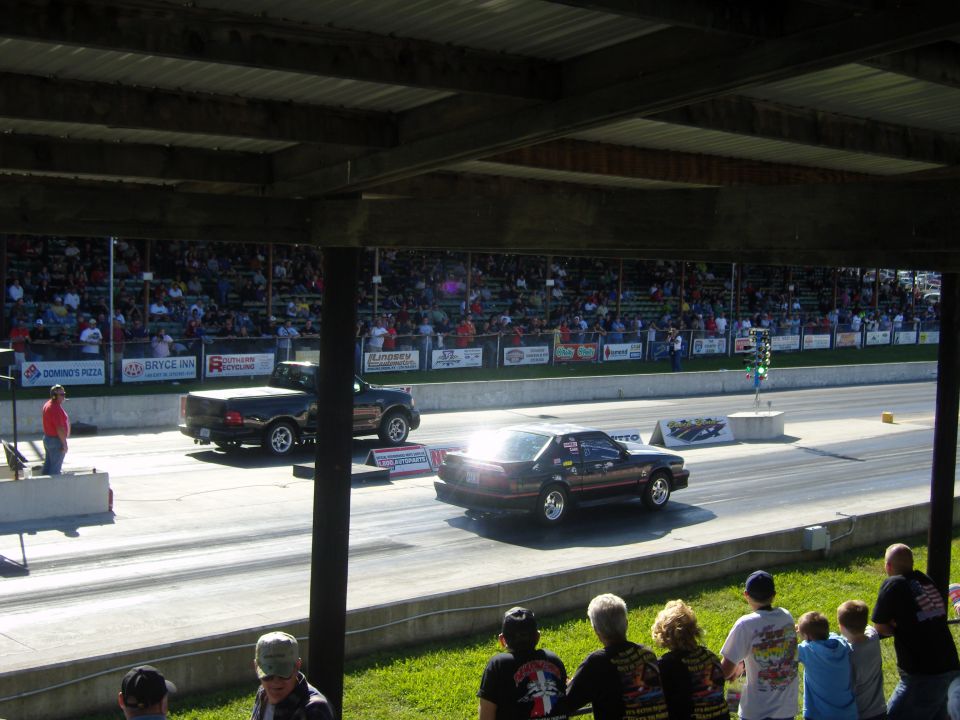 Two cars racing on a drag strip with spectators in the background