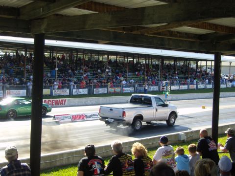 2007 Ford Mustang - A green car and a silver pickup truck compete on a drag strip, with a crowd watching from the bleachers.