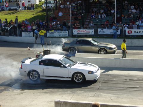 2007 Ford Mustang - Two Ford Mustangs prepare for a drag race, with the white car kicking up smoke from its tires.